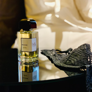 A fragrance sits on a hotel nightstand beside a bra