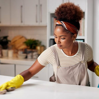 Young woman wearing yellow gloves cleaning a kitchen countertop with a cloth and spray bottle, practicing odor-neutralizing techniques for a fresh-smelling home