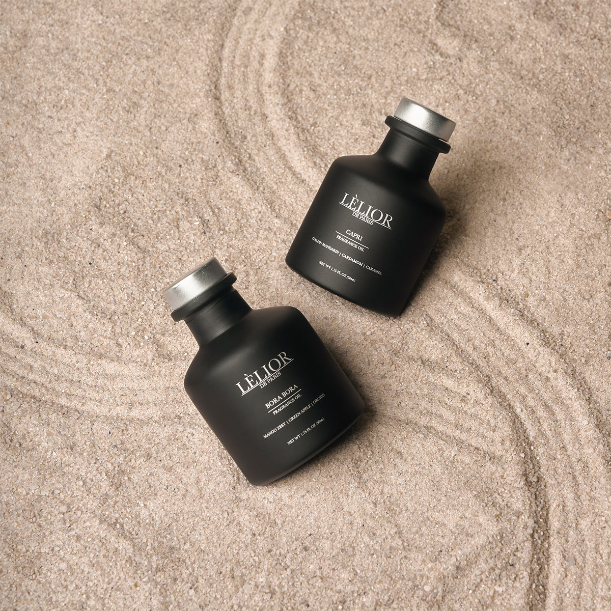 2 black 50ml bottles with silver caps and silver lettering. Bottles laying flat on the sand. Bora Bora and Capri home fragrance oils. 