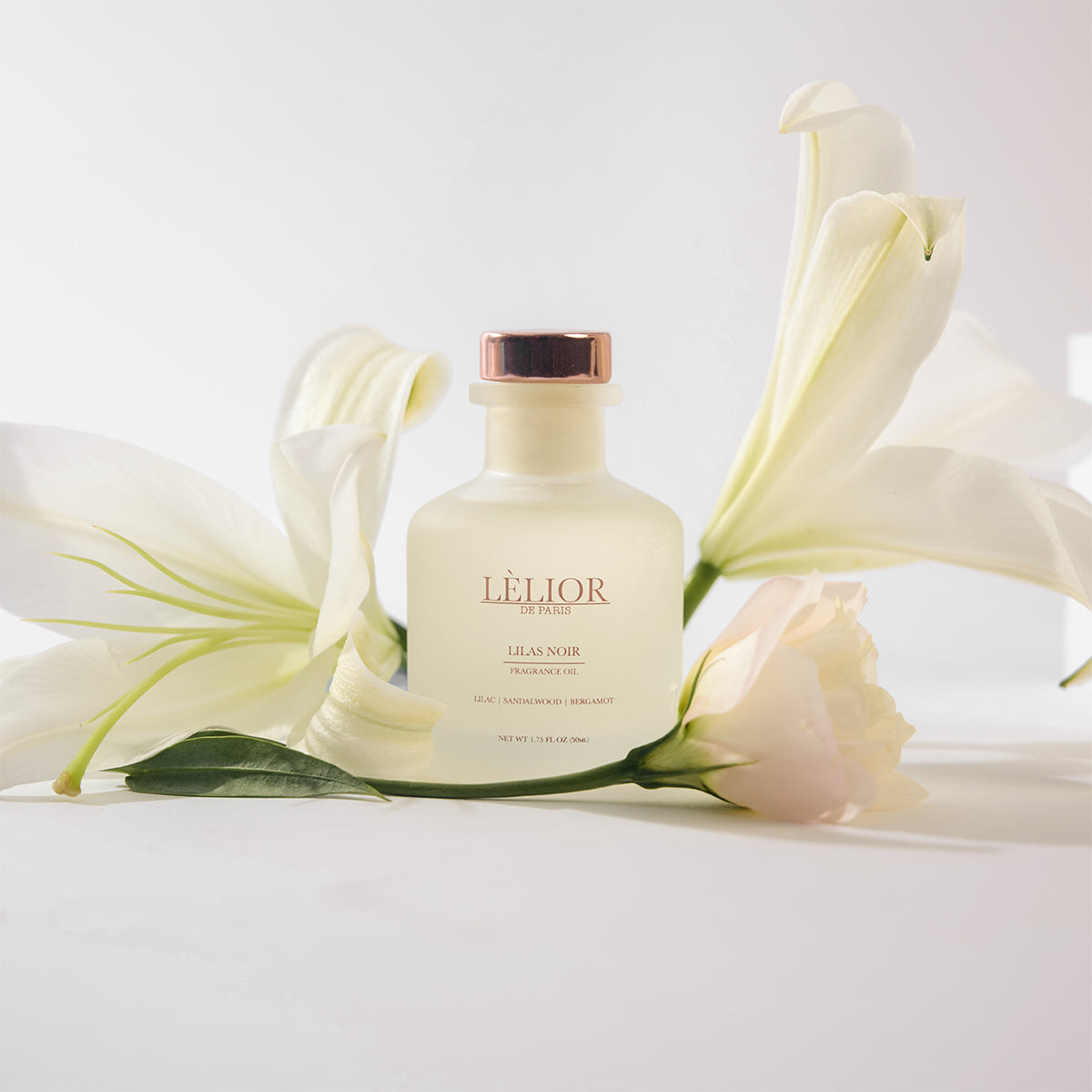 Elegant 50ml bottle of Lilas Noir fragrance oil with beautiful flowers surrounding the bottle. Frost glass bottle with rose gold lettering and cap. On the bottle, notes of lilac, sandalwood, and bergamot are described. White background. 